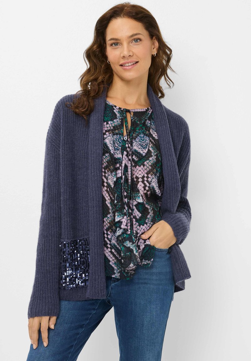 Cardigan en tricot bleu marine avec une finition texturée et une poche à sequins, superposé à une blouse à motifs dans des teintes de vert et de violet, associée à un jean bleu.
