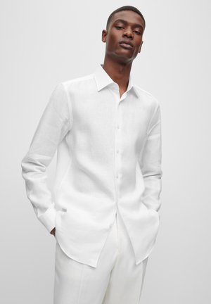 White linen shirt with long sleeves and a pointed collar, featuring a button-down front and curved hem. Smooth texture, minimal detailing.