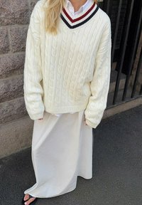 Suéter crema de punto trenzado con un escote en V profundo, adornado con rayas azul marino y rojas. Combinado con una falda larga y lisa blanca y sandalias negras.