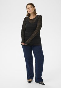 Femme se tenant debout, portant un haut en dentelle noir à manches longues, un jean large bleu foncé et des escarpins noirs à bout pointu sur un fond blanc.