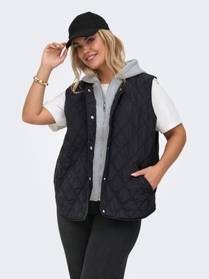 Smilende kvinne med svart caps, svart quiltet vest over grå glidelåshoodie, hvit t-skjorte, svarte bukser, med den ene hånden i lommen og den andre som justerer capsen.