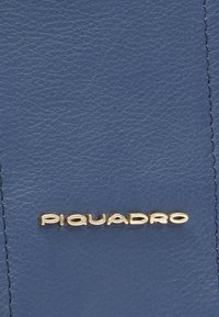 Pelle blu texturizzata con logo "Piquadro" dorato. Presenta cuciture visibili lungo i bordi, che aggiungono dettagli al design.