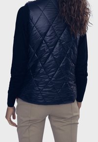 Tmavě modrá prošívaná péřová vesta s lesklým povrchem, kombinovaná s černým topem s dlouhým rukávem a béžovými kalhotami. Viditelný texturovaný diamantový vzor.