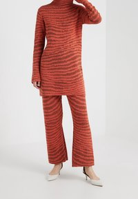 Personne portant une robe-pull tricotée orange-rouge et un pantalon assorti avec des rayures noires ondulées, associée à des escarpins blancs à bouts pointus.