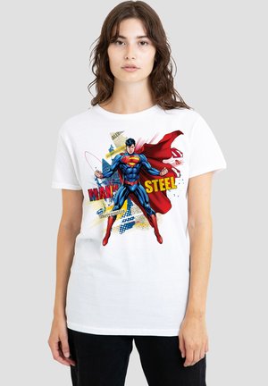 Woman wearing a white t-shirt with a colorful Superman graphic and red cape, standing against a plain light gray background.