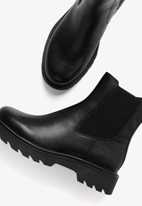 Mustad musta nahast pahkluu saapad elastsete külgpaneelidega, tekstuuriga viimistluse, ümarate varvastega ning kareda soledega, mis on varustatud sügavate soontega.