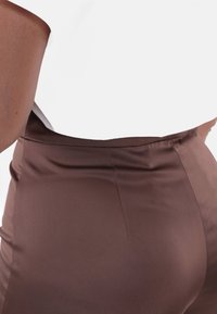 Pantaloni sartoriali marroni in satin con una texture liscia, dotati di vita alta e design elegante. Dettaglio cuciture visibile sul retro.