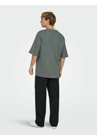 Grijs katoenen T-shirt met een losse pasvorm, korte mouwen en een ronde hals, gecombineerd met zwarte broek en witte sneakers. Gefotografeerd van achteren.