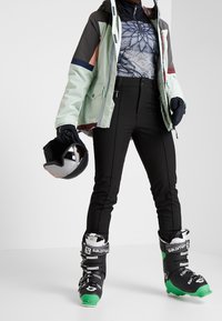 Strój narciarski z wielokolorową kurtką, wzorzystą termiczną bluzą, czarnymi spodniami i czarnymi butami narciarskimi z zielonymi akcentami; w ręku kask.