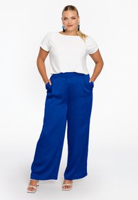 Weißes kurzärmeliges Oberteil, kombiniert mit lebhaft blauen, weit geschnittenen Hosen mit Seitentaschen. Frau trägt minimalistische weiße Absätze und Ohrringe.