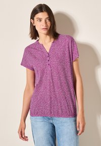 Halenka s krátkým rukávem v purpurové barvě se geometrickým vzorem, výstřihem do V a čtyřmi knoflíky. Měkká tkanina s volným střihem.