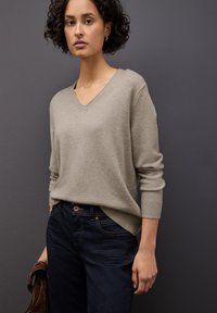 Suéter de pico beige claro con puños acanalados, combinado con jeans azul oscuro. Textura suave con un ajuste relajado, llevado de manera casual.