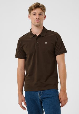 Joven con el pelo claro que lleva una camisa polo marrón oscuro y vaqueros azules, de pie con una expresión neutra contra un fondo liso.