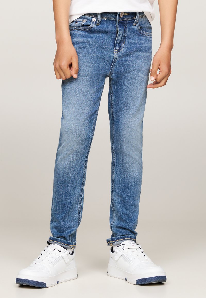 Plave traperice uskog kroja s laganim izblijedjenjem, uparene s bijelim tenisicama s tamnoplavim detaljima i teksturiranom potplatkom.