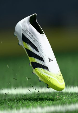 White soccer cleat with black stripes and yellow accents, textured upper and a low-profile design. Elevated above green grass.