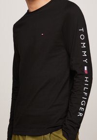 Schwarzes Langarmshirt mit kleinem Logo auf der Brust und "TOMMY HILFIGER"-Text entlang des linken Ärmels. Baumwollmaterial, taillierter Schnitt.