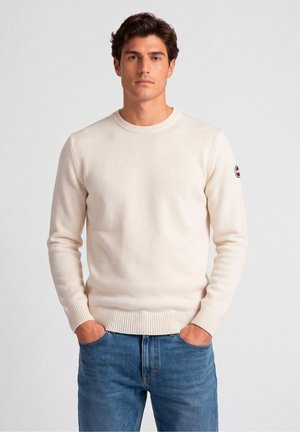 Uomo che indossa un maglione lavorato a maglia color crema con un logo sulla manica e jeans blu di denim, in piedi con le mani in tasca.