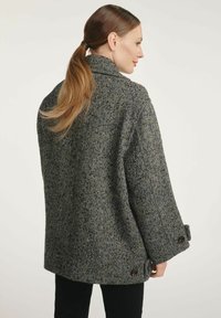 Kobieta z jasnobrązowym kucykiem, ubrana w szary płaszcz w jodełkę z zapiętymi mankietami, stoi na tle jasnego, jednolitego tła.