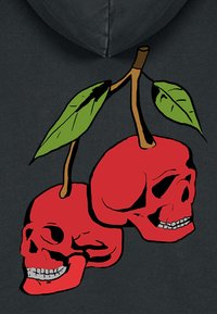 Grafika červených lebiek s čiernym obrysom a zelenými listami, navrhnutá na tmavom mikinovom svetri s kapucňou. Lebky sú naskladané s hnedým stonkom.
