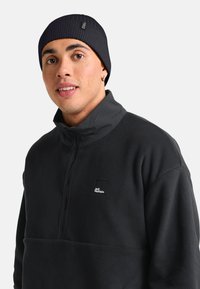 Giovane uomo che indossa un berretto nero a coste e una giacca in fleece nera Jack Wolfskin con collo a mezza zip, che guarda leggermente di lato.