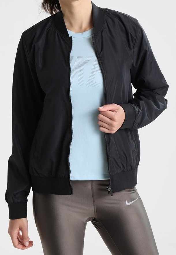 Femme portant une veste noire zippée sur un t-shirt bleu clair avec un imprimé graphique et un legging gris Nike.