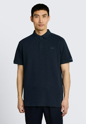 Jeune homme portant un polo uni bleu marine foncé à manches courtes, debout devant un fond gris clair.