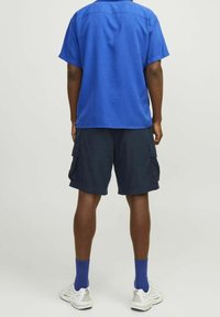 Camicetta blu a maniche corte, pantaloni cargo blu scuro con tasche laterali, sneakers bianche e calzini blu. Outfit casual visto da dietro.