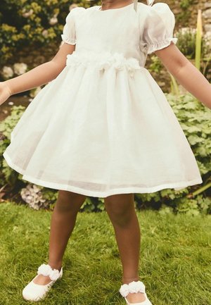 Robe en lin blanc avec des manches bouffantes et un détail floral à la taille, associée à des chaussures blanches ornées d'accents floraux. Jupe courte et ample.