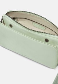 Grüne Ledertasche mit Umhängegurt und Reißverschluss, mit einer glatten Außenseite und einem kontrastierenden hellen Innenraum. Enthält ein Markenlabel im Inneren.