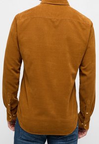 Camicia in corduroy marrone con colletto, maniche lunghe e polsini con bottoni. Presenta una texture liscia e un orlo curvo. Colore uniforme su tutta la superficie.
