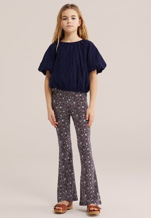 Jauna mergina stovinti, dėvinti tamsiai mėlyną palaidinę su pūstomis rankovėmis, gėlių raštuotus platėjančius kelnes ir rudus sandalus neutralaus fono fone.