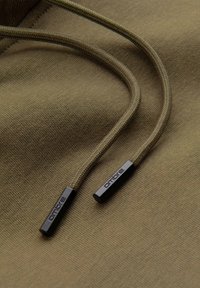 Nahaufnahme von olivgrünem Hoodie-Stoff mit passenden Zugbändern, die in schwarzen Kunststoffenden mit der Aufschrift "ombre" enden.