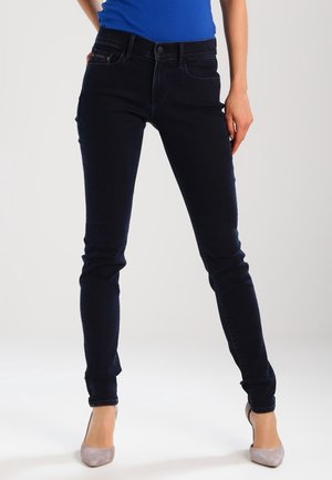 Vrouw draagt donkerblauwe skinny jeans, een blauwe top en lichtgrijze hoge hakken, staand met gespreide benen op een witte achtergrond.