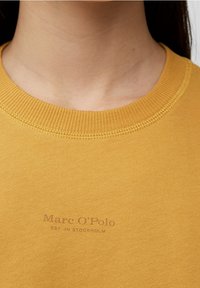 Sudadera amarilla con escote acanalado, que presenta un sutil logotipo impreso "Marc O'Polo EST. EN ESTOCOLMO" en la parte frontal. Textura suave.