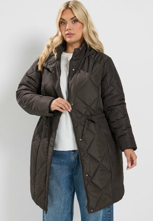 Femme portant un long manteau matelassé noir avec une ceinture à la taille, par-dessus un pull blanc et un jean bleu, debout devant un fond clair uni.