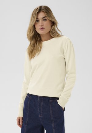 Žena s dlhými blond vlasmi, oblečená v krémovom svetri s dlhým rukávom a tmavomodrých nohaviciach s vysokým pásom s detailmi švov, stojaca čelom vpred.