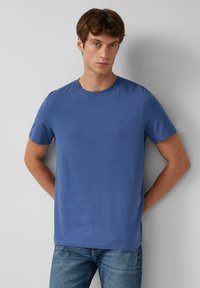 Blaues Baumwoll-T-Shirt mit kurzen Ärmeln, Rundhalsausschnitt und dezentem Branding auf der Brust. Kombiniert mit Jeans. Glatte Textur.