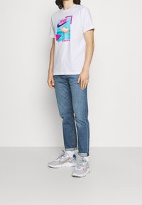 Vit t-shirt med kort ärm som har en färgglad flamingografik, tillsammans med ljusblå jeans och grå Nike-sneakers med röda detaljer.