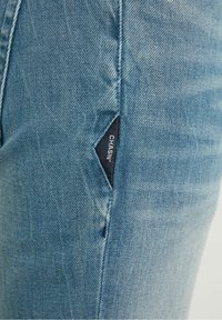 Jeans de mezclilla azul claro con una pequeña etiqueta de logo negra en un corte en el muslo, con un acabado texturizado y ligeramente desgastado.
