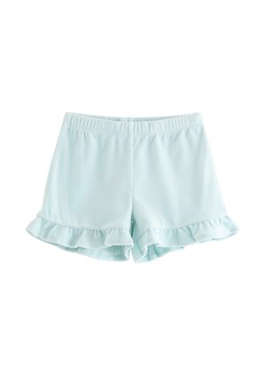 Smilende ung pige, der står med hænderne ned langs siderne, iført en cremefarvet skjorte med flæser på ærmerne og papegøjedesign, lyseblå flæse-short og farverige sneakers.