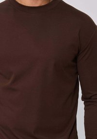 Chemise marron à manches longues en tissu doux, avec un col rond et une coupe ajustée, caractérisée par une texture lisse et un design minimaliste.