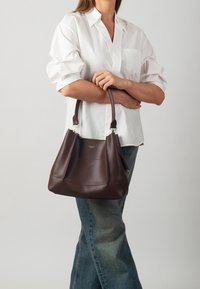 Bolso de mano de cuero marrón con forma estructurada, que cuenta con dos asas y detalles sutiles de costura. Combinado con una camisa blanca y jeans de mezclilla.