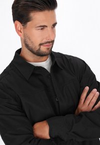 Camicia nera con bottone, caratterizzata da colletto, maniche lunghe e una texture liscia. Design minimalista con dettagli di cucitura visibili.