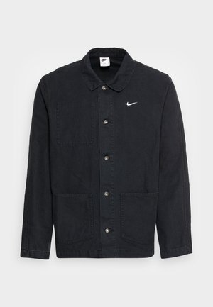 Chaqueta de algodón negra con cuello, cierre frontal de botones, dos bolsillos de parche y un pequeño logotipo blanco de Nike en el lado izquierdo del pecho.