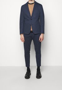 Blazer navy con due bottoni e tasca, abbinato a un maglione girocollo marrone e pantaloni tapered coordinati. Mocassini neri completano l'outfit.