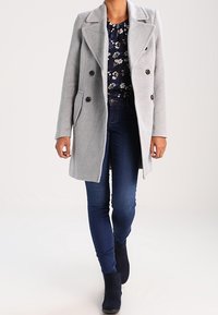 Cappotto grigio doppiopetto con ampi revers, tasche laterali e bottoni scuri, indossato sopra un top floreale blu e jeans scuri.