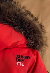 Červená bunda s textúrovanou hnedou kožušinovou kapucňou. Obsahuje vyšívané logo "SUPER DRY JPN." v čiernej a bielej farbe na ľavej strane hrude.