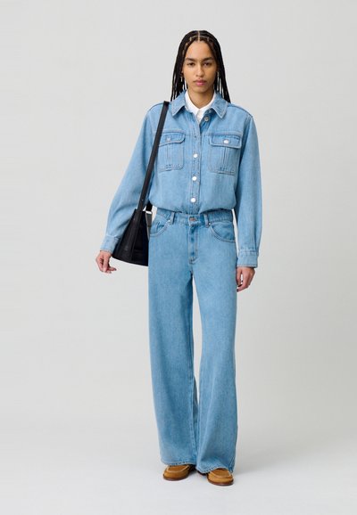 Camicia in denim blu chiaro con due taschini sul petto e chiusura a bottoni, abbinata a pantaloni in denim blu chiaro a gamba larga e scarpe marroni slip-on.