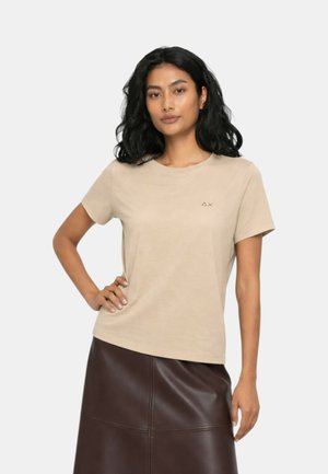 Donna con capelli neri ondulati, che indossa una maglietta beige a maniche corte e una gonna in pelle marrone scuro, in piedi con una mano sul fianco.