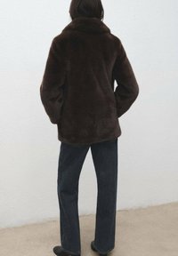 Bruine jas van imitatiebont met een hoge kraag, losse pasvorm en lange mouwen; gecombineerd met donkere jeans met rechte pijpen. Neutrale achtergrond.